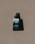 Ninth Wave First Point Daily Facial Cleanser bottle placed on the sand at the beach to reflect a post-surf cleanser. 